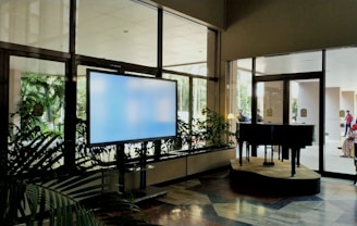 A spacious indoor area featuring a large digital screen on the left, surrounded by potted plants. To the right, a grand piano is positioned on a small stage near large glass doors. Sunlight illuminates the scene, creating reflections on the glass surfaces. People are visible outside beyond the doors, indicating a busy and active environment.