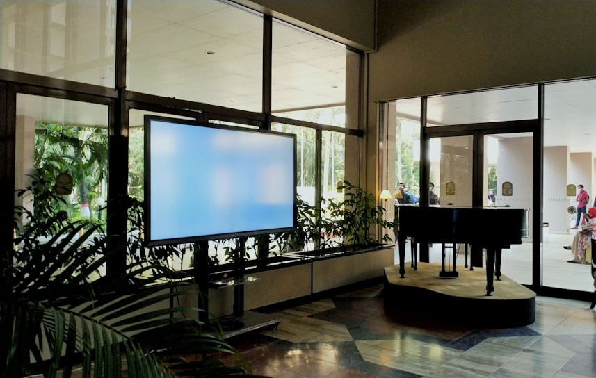 A spacious indoor area featuring a large digital screen on the left, surrounded by potted plants. To the right, a grand piano is positioned on a small stage near large glass doors. Sunlight illuminates the scene, creating reflections on the glass surfaces. People are visible outside beyond the doors, indicating a busy and active environment.