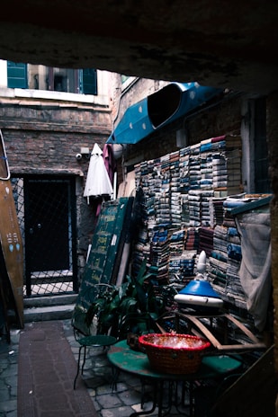 An alleyway with stacks of books lined against a brick wall. There are several green metal chairs and a table with a basket on it. A blue awning partially shelters the area. A door with a metal grate leads to a building, and plants are placed near the entrance.