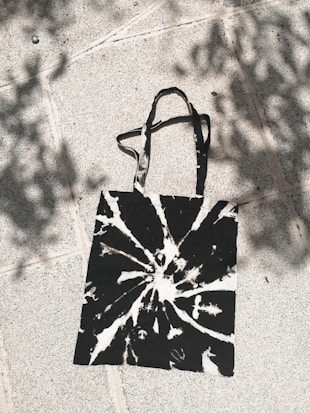 A tie-dye tote bag with black and white patterns lies on a sunlit, textured pavement. Shadows of leaves create an artistic play of light and shadow on the surface.