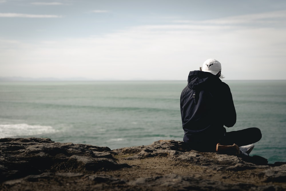 person in black jacket sitting on brown rock near body of water during daytime