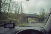 Raindrops on a car windshield with a view of a road and bridge outside. The windshield has a GPS device mounted on the dashboard, displaying navigation directions. A car is visible in front, driving down a wet road surrounded by trees.