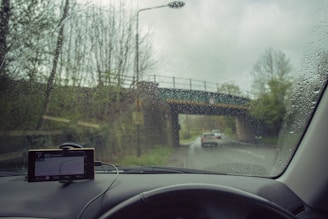 Raindrops on a car windshield with a view of a road and bridge outside. The windshield has a GPS device mounted on the dashboard, displaying navigation directions. A car is visible in front, driving down a wet road surrounded by trees.