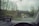 Raindrops on a car windshield with a view of a road and bridge outside. The windshield has a GPS device mounted on the dashboard, displaying navigation directions. A car is visible in front, driving down a wet road surrounded by trees.