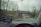 Raindrops on a car windshield with a view of a road and bridge outside. The windshield has a GPS device mounted on the dashboard, displaying navigation directions. A car is visible in front, driving down a wet road surrounded by trees.