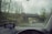 Raindrops on a car windshield with a view of a road and bridge outside. The windshield has a GPS device mounted on the dashboard, displaying navigation directions. A car is visible in front, driving down a wet road surrounded by trees.