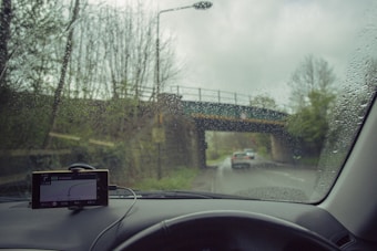 Raindrops on a car windshield with a view of a road and bridge outside. The windshield has a GPS device mounted on the dashboard, displaying navigation directions. A car is visible in front, driving down a wet road surrounded by trees.