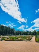 A well-manicured garden features geometric hedges, colorful flower beds, and classical statues under a vibrant blue sky with scattered clouds. The garden paths are neatly aligned, and the lush trees form a dense backdrop.