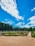 A well-manicured garden features geometric hedges, colorful flower beds, and classical statues under a vibrant blue sky with scattered clouds. The garden paths are neatly aligned, and the lush trees form a dense backdrop.