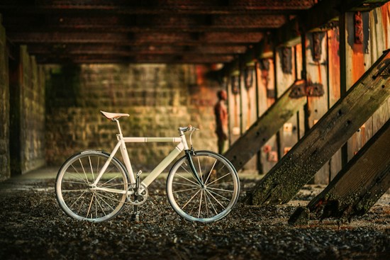 A sleek, modern bicycle is positioned against a rustic, textured backdrop of large wooden beams and weathered concrete walls under a dimly lit structure. The light softly illuminates the bicycle&rsquo;s metallic frame, highlighting its minimalist design.