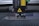 A laser engraving or cutting machine is positioned above a metallic surface, with its nozzle directed downward. The machine features warning labels, and the environment appears industrial, with a grid-like surface underneath.