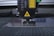 A laser engraving or cutting machine is positioned above a metallic surface, with its nozzle directed downward. The machine features warning labels, and the environment appears industrial, with a grid-like surface underneath.