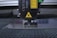 A laser engraving or cutting machine is positioned above a metallic surface, with its nozzle directed downward. The machine features warning labels, and the environment appears industrial, with a grid-like surface underneath.