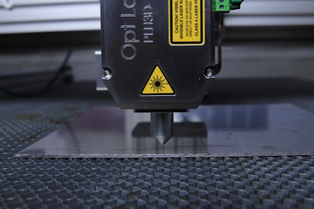 A laser engraving or cutting machine is positioned above a metallic surface, with its nozzle directed downward. The machine features warning labels, and the environment appears industrial, with a grid-like surface underneath.