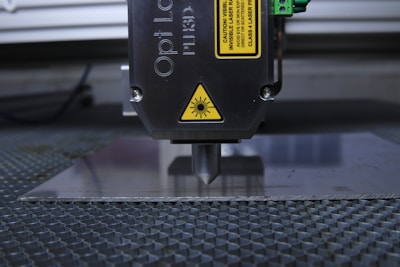 A laser engraving or cutting machine is positioned above a metallic surface, with its nozzle directed downward. The machine features warning labels, and the environment appears industrial, with a grid-like surface underneath.