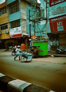 A man wearing a conical hat is pushing a cart filled with metal containers along a street. There are motorcycles parked nearby, and a green vending stall is visible near the sidewalk. The surroundings include buildings with commercial signs and many electrical wires overhead.