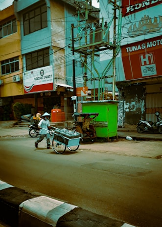 A man wearing a conical hat is pushing a cart filled with metal containers along a street. There are motorcycles parked nearby, and a green vending stall is visible near the sidewalk. The surroundings include buildings with commercial signs and many electrical wires overhead.