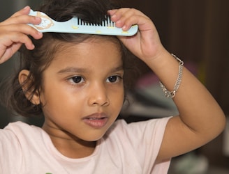 A child wearing a light-colored shirt is using a blue comb with cartoon designs to brush their hair. The child is focused and has a bracelet on one wrist, with a soft background out of focus.