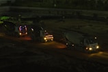 Three cement mixer trucks are parked on a construction site at nighttime. They are lined up on a dirt path, illuminated by their own headlights and some ambient light, casting shadows in the surrounding area. The background features a dim construction area with few visible details.