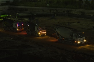 Three cement mixer trucks are parked on a construction site at nighttime. They are lined up on a dirt path, illuminated by their own headlights and some ambient light, casting shadows in the surrounding area. The background features a dim construction area with few visible details.