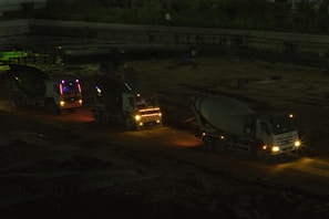 Three cement mixer trucks are parked on a construction site at nighttime. They are lined up on a dirt path, illuminated by their own headlights and some ambient light, casting shadows in the surrounding area. The background features a dim construction area with few visible details.