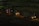 Three cement mixer trucks are parked on a construction site at nighttime. They are lined up on a dirt path, illuminated by their own headlights and some ambient light, casting shadows in the surrounding area. The background features a dim construction area with few visible details.