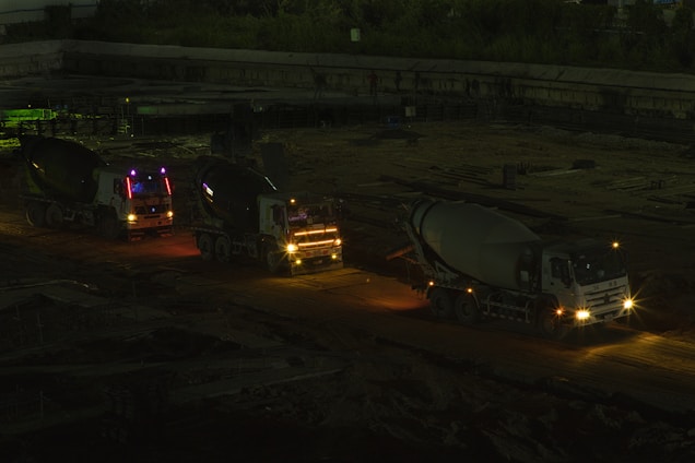 Three cement mixer trucks are parked on a construction site at nighttime. They are lined up on a dirt path, illuminated by their own headlights and some ambient light, casting shadows in the surrounding area. The background features a dim construction area with few visible details.