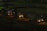 Three cement mixer trucks are parked on a construction site at nighttime. They are lined up on a dirt path, illuminated by their own headlights and some ambient light, casting shadows in the surrounding area. The background features a dim construction area with few visible details.