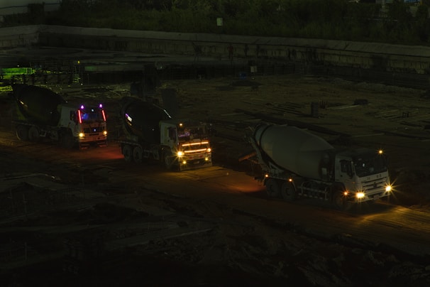Three cement mixer trucks are parked on a construction site at nighttime. They are lined up on a dirt path, illuminated by their own headlights and some ambient light, casting shadows in the surrounding area. The background features a dim construction area with few visible details.