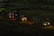 Three cement mixer trucks are parked on a construction site at nighttime. They are lined up on a dirt path, illuminated by their own headlights and some ambient light, casting shadows in the surrounding area. The background features a dim construction area with few visible details.