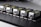 Five identical industrial components are aligned in a row on a perforated black surface. Each device has a metallic finish with labels and warning signs visible. The components are compact and connected through visible cables.
