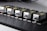 Five identical industrial components are aligned in a row on a perforated black surface. Each device has a metallic finish with labels and warning signs visible. The components are compact and connected through visible cables.