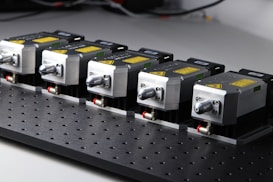 Five identical industrial components are aligned in a row on a perforated black surface. Each device has a metallic finish with labels and warning signs visible. The components are compact and connected through visible cables.