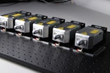 Five identical metallic devices are aligned in a row on a perforated black surface. Each device features warning labels and has a compact, industrial design, suggesting they are precision instruments or components used in an engineering or laboratory setting.