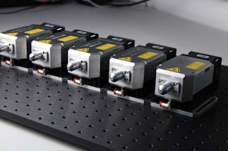 Five identical metallic devices are aligned in a row on a perforated black surface. Each device features warning labels and has a compact, industrial design, suggesting they are precision instruments or components used in an engineering or laboratory setting.