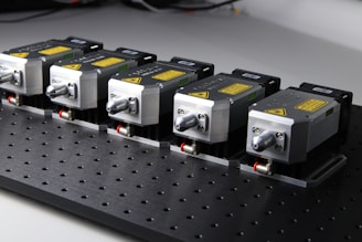 Five identical metallic devices are aligned in a row on a perforated black surface. Each device features warning labels and has a compact, industrial design, suggesting they are precision instruments or components used in an engineering or laboratory setting.