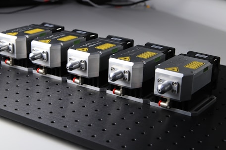 Five identical metallic devices are aligned in a row on a perforated black surface. Each device features warning labels and has a compact, industrial design, suggesting they are precision instruments or components used in an engineering or laboratory setting.