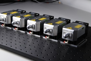 Five identical metallic devices are aligned in a row on a perforated black surface. Each device features warning labels and has a compact, industrial design, suggesting they are precision instruments or components used in an engineering or laboratory setting.