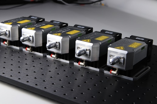 Five identical metallic devices are aligned in a row on a perforated black surface. Each device features warning labels and has a compact, industrial design, suggesting they are precision instruments or components used in an engineering or laboratory setting.