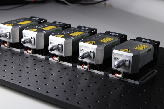 Five identical metallic devices are aligned in a row on a perforated black surface. Each device features warning labels and has a compact, industrial design, suggesting they are precision instruments or components used in an engineering or laboratory setting.