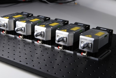 Five identical metallic devices are aligned in a row on a perforated black surface. Each device features warning labels and has a compact, industrial design, suggesting they are precision instruments or components used in an engineering or laboratory setting.