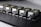 Five identical metallic devices are aligned in a row on a perforated black surface. Each device features warning labels and has a compact, industrial design, suggesting they are precision instruments or components used in an engineering or laboratory setting.