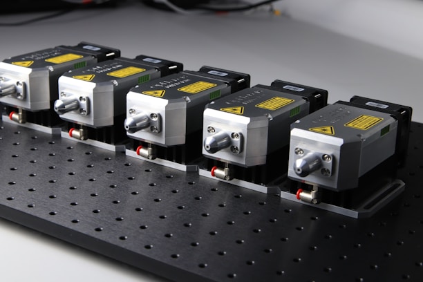 Five identical metallic devices are aligned in a row on a perforated black surface. Each device features warning labels and has a compact, industrial design, suggesting they are precision instruments or components used in an engineering or laboratory setting.