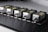 Five identical metallic devices are aligned in a row on a perforated black surface. Each device features warning labels and has a compact, industrial design, suggesting they are precision instruments or components used in an engineering or laboratory setting.
