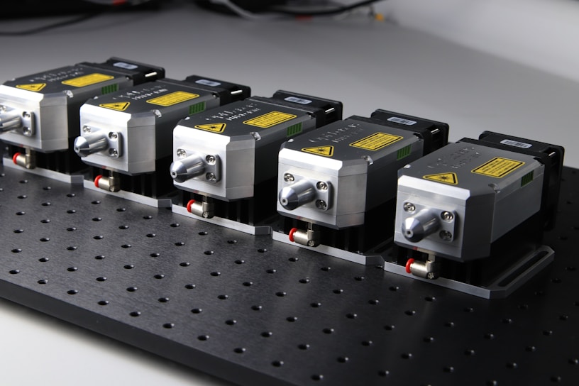 Five identical metallic devices are aligned in a row on a perforated black surface. Each device features warning labels and has a compact, industrial design, suggesting they are precision instruments or components used in an engineering or laboratory setting.
