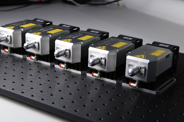 Five identical metallic devices are aligned in a row on a perforated black surface. Each device features warning labels and has a compact, industrial design, suggesting they are precision instruments or components used in an engineering or laboratory setting.