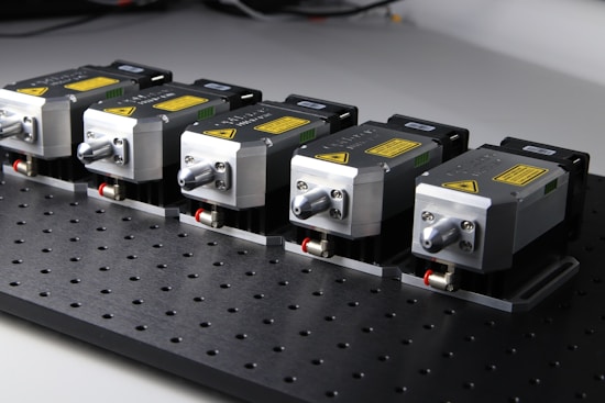 Five identical metallic devices are aligned in a row on a perforated black surface. Each device features warning labels and has a compact, industrial design, suggesting they are precision instruments or components used in an engineering or laboratory setting.