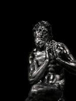 A detailed and realistic sculpture of a seated male figure with notable muscular definition. The material has a dark metallic sheen, reflecting light and emphasizing the contours of the figure's body. The figure has a contemplative expression, with his hands clasped together near his face.