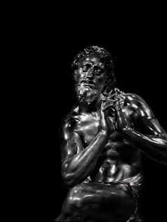 A detailed and realistic sculpture of a seated male figure with notable muscular definition. The material has a dark metallic sheen, reflecting light and emphasizing the contours of the figure's body. The figure has a contemplative expression, with his hands clasped together near his face.