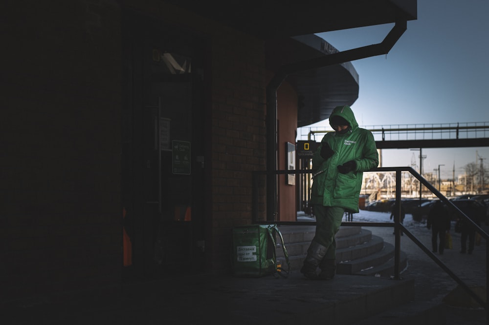 緑色のジャケットと緑のズボンを着た男が、夜中に茶色の建物の近くに立っている